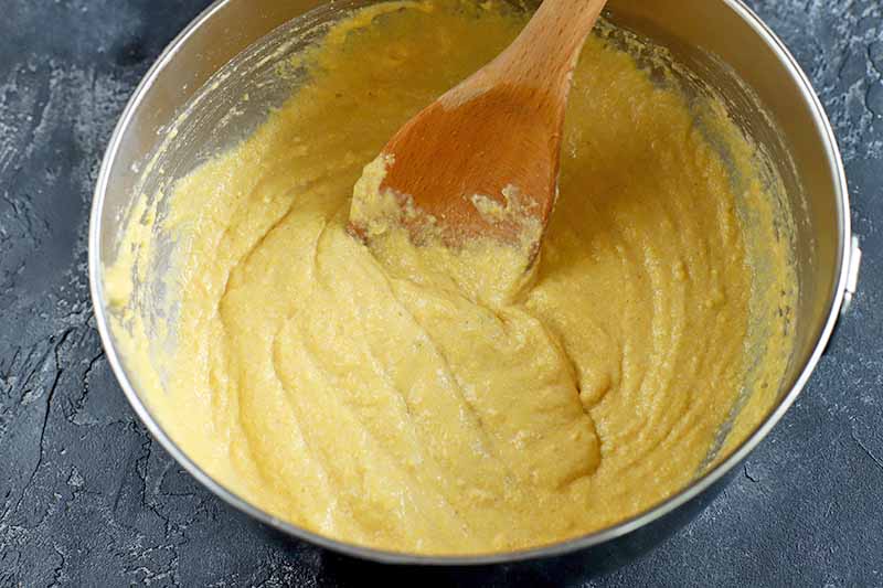 Overhead closely cropped horizontal image of yellow cornbread batter in a stainless steel mixing bowl, being stirred with a wooden spoon, on a gray stone surface.