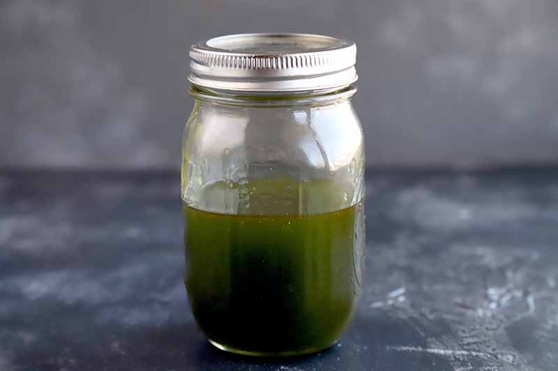Green flavored oil fills a glass jar about 2/3 of the way to the top, with a metal lid, on a marked gray stone surface, against a lighter gray background.