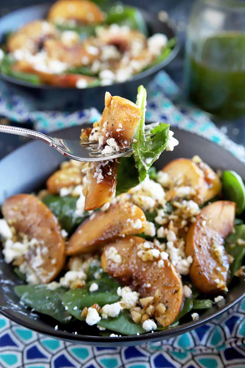 Vertical image of a forkful of peach and spinach salad being held up to the camera, with more in two gray bowls in the background, on a dark and light blue patterned cloth, with a jar of homemade basil-infused olive oil to the right.
