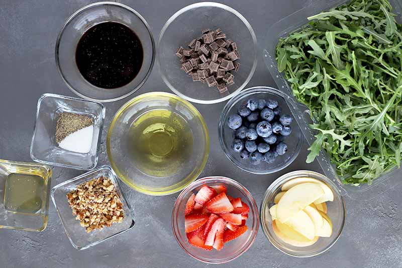 Overhead image of three small square and six medium round glass bowls of oil, honey, salt and pepper, chopped valnuts, chocolate chips, balsamic vinegar, chopped strawberries, whole blueberries, and sliced apples, with a pile of fresh arugula to the right, on a gray surface.