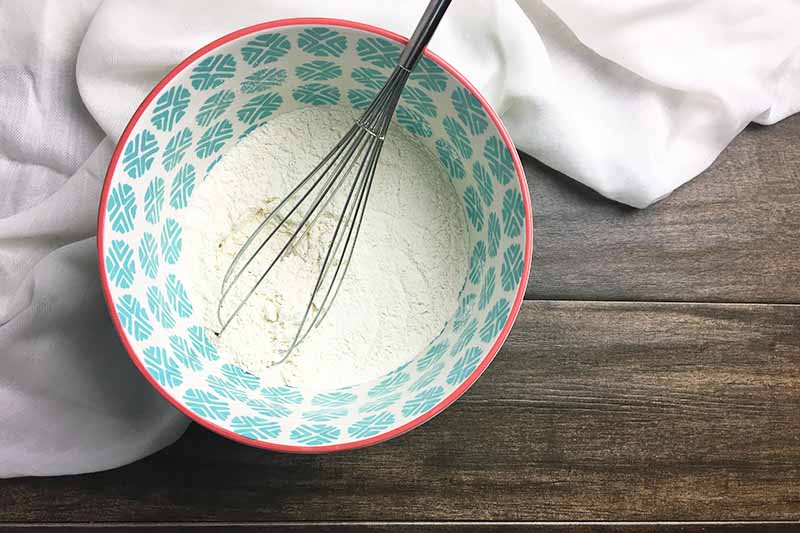 Horizontal image of a whisk in a blue bowl with dry ingredients on a white towel.
