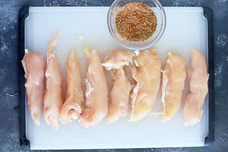 Horizontal image of a cutting board with uncooked poultry tenders and a bowl of mixed spices.