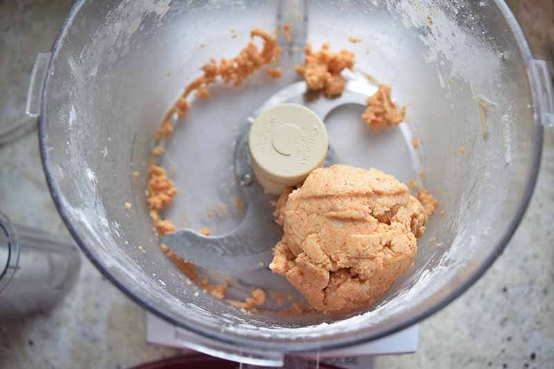 An orange dough ball and crumbs in the bottom of a food processor on a gray speckled kitchen countertop.