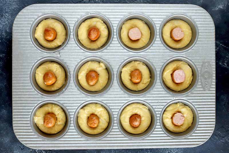 Overhead tightly framed image of a metal muffin tin filled with cornbread batter, with a piece of hot dog centered in each, on a dark gray surface.