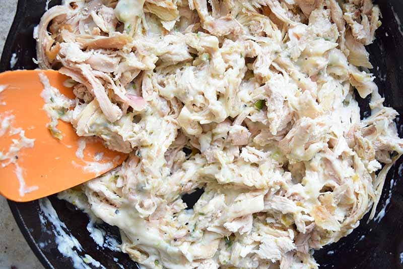 Closeup horizontal image of shredded chicken in a creamy sauce in a large frying pan, being stirred with an orange spatula.