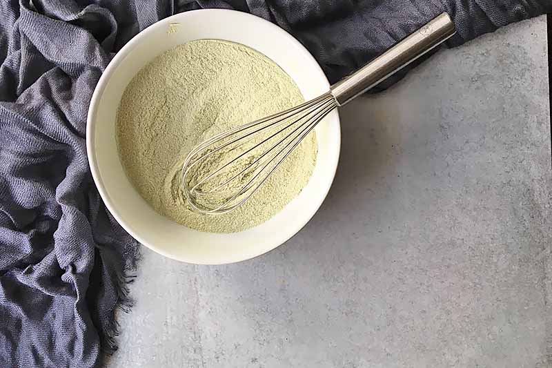 Horizontal image of a light green dry mixture in a white bowl stirred with a whisk.
