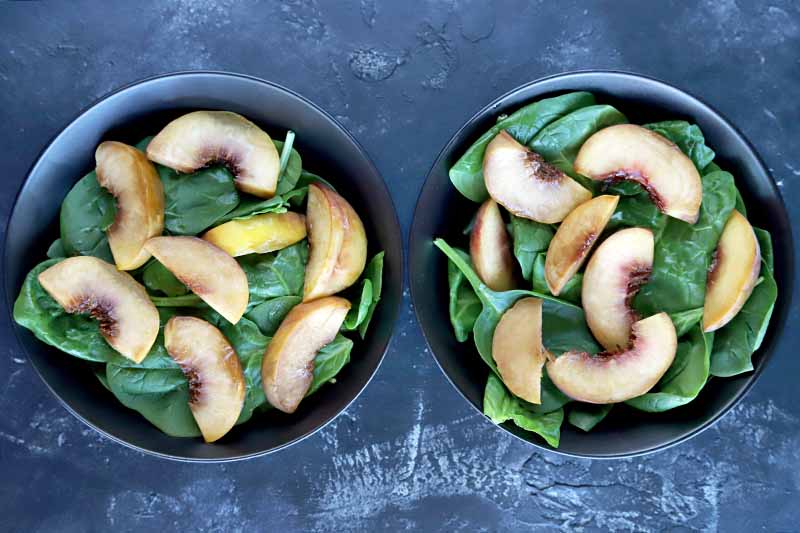 Overhead horizontal image of two gray bowls filled with raw baby spinach, with honey-glazed roasted peach slices with the skin on arranged on top, on a gray background.