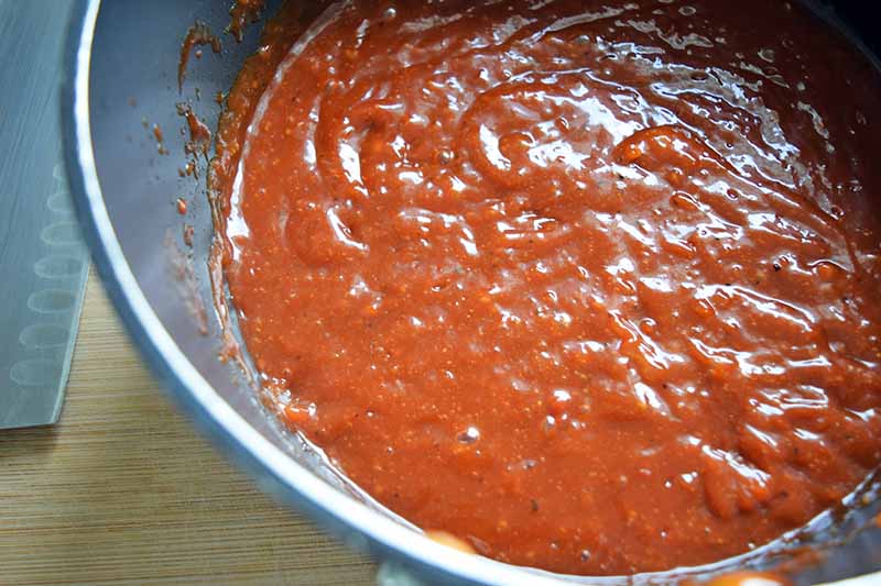 Closely cropped overhead horizontal image of a pan of red sauce, on a beige surface with the blade of a chef's knife to the left.