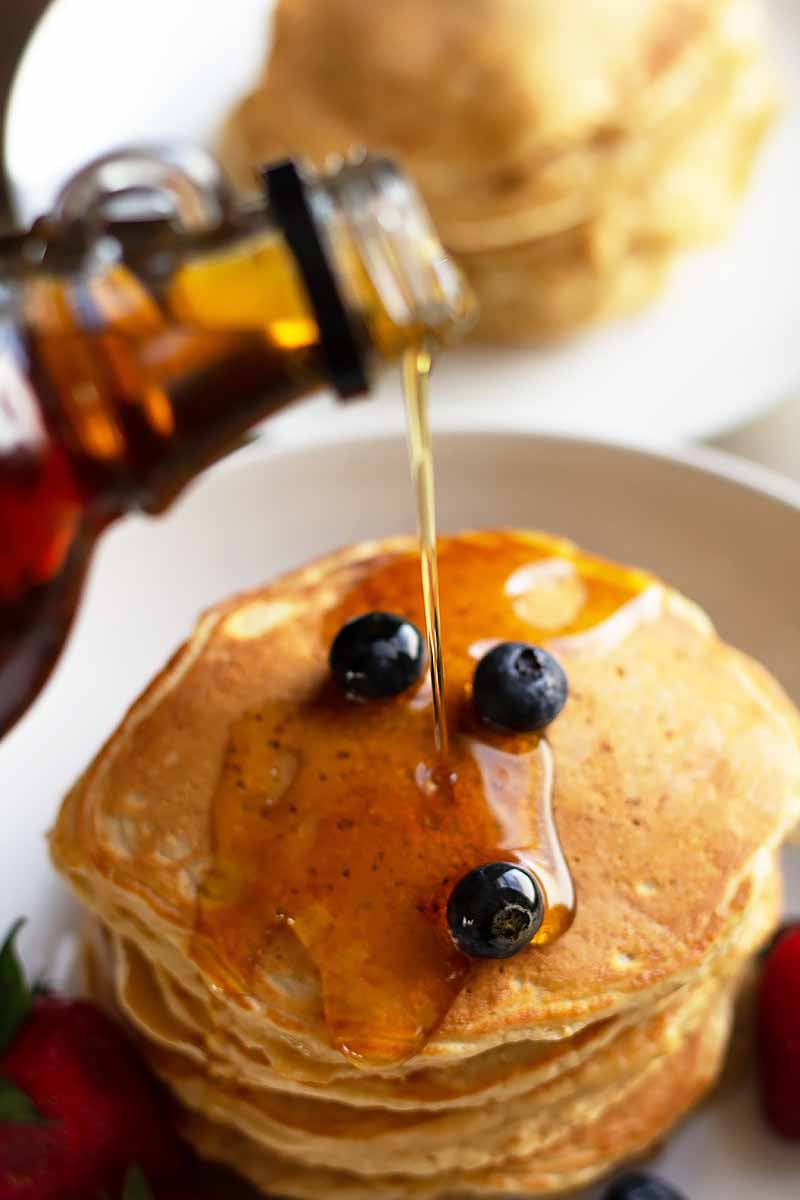 Vertical image of maple syrup pouring over a stack of flapjacks topped with three blueberries.