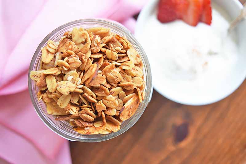 Horizontal image of a glass of granola and a bowl of yogurt on a wooden table with a pink towel.