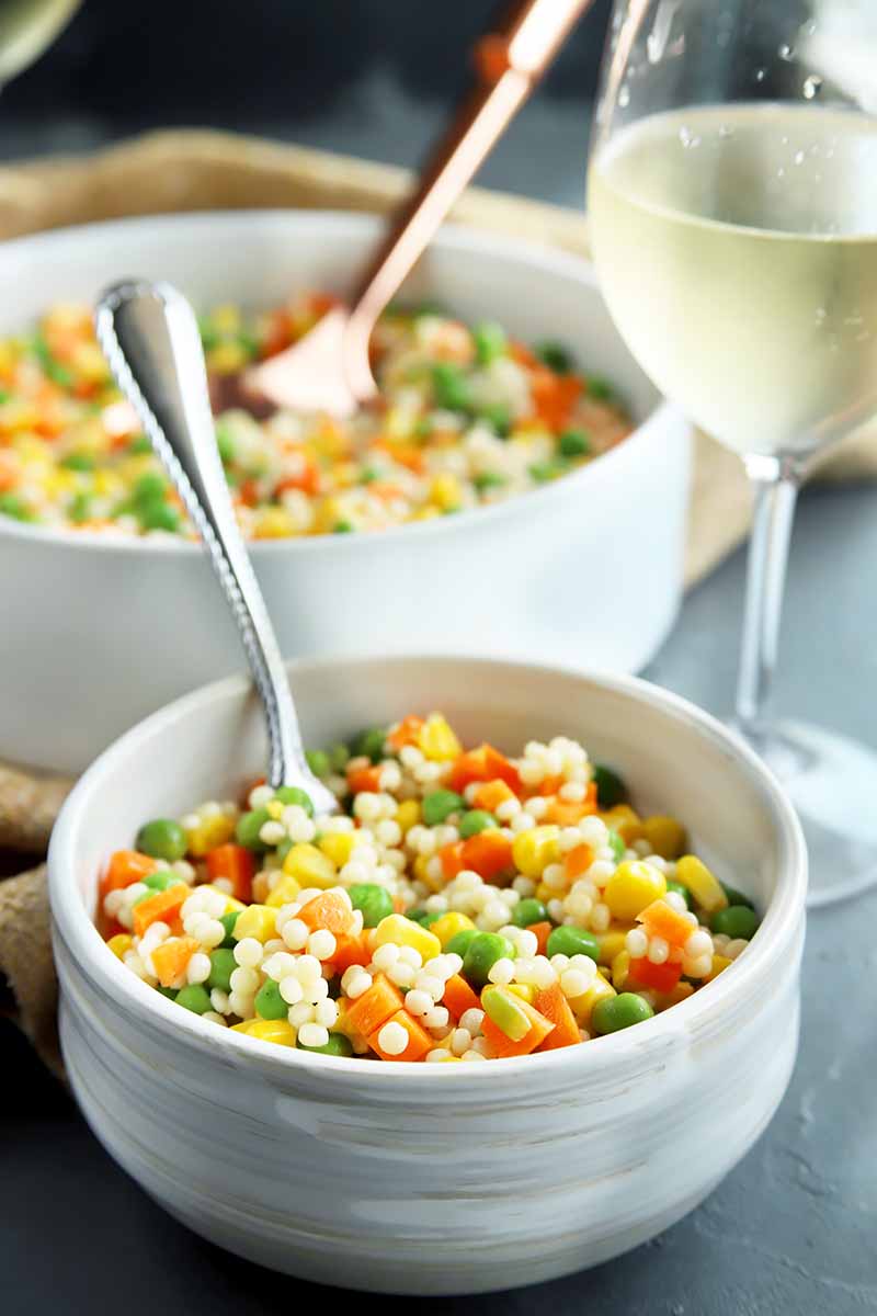 Vertical image of two white bowls with a veggie and grain mix and spoons inserted into each spoon next to a glass of white wine.