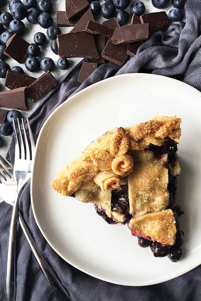 Vertical top-down image of a white plate with a slice of pie, next to blueberries, chocolate chunks, and forks.