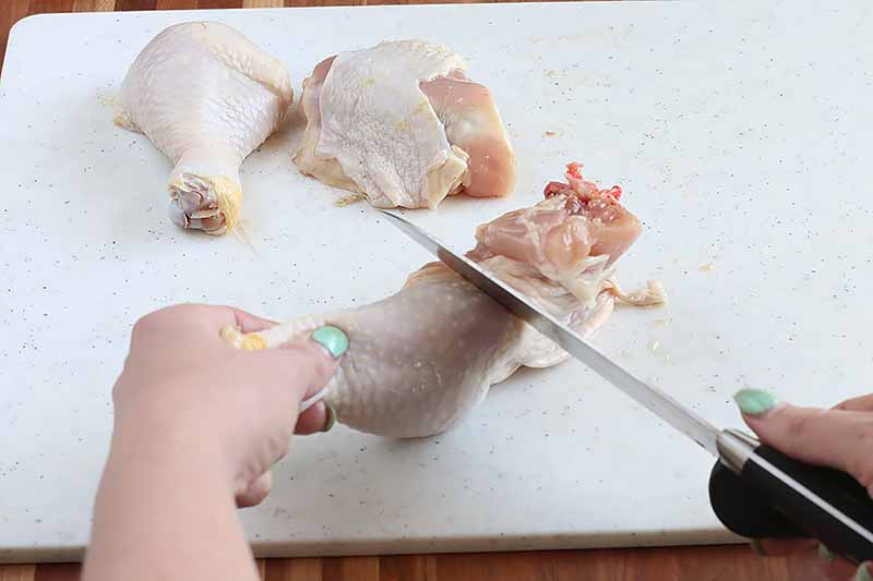 Horizontal image of a hand holding a piece of raw chicken while a knife cuts through it, with a drumstick and thigh in the background on a white cutting board.