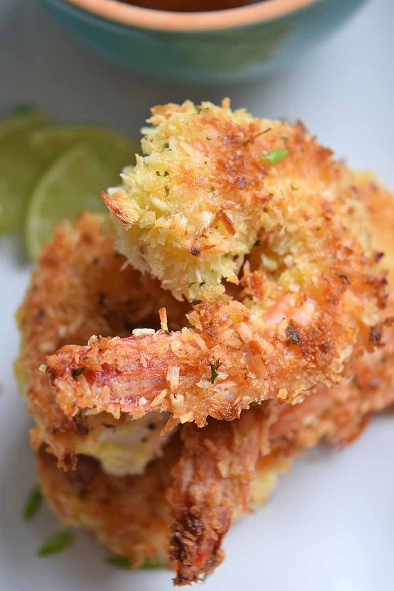 Vertical overhead image of a small stack of baked coconut shrimp, with slices of lime and small blue bowl of sauce, on a white plate.