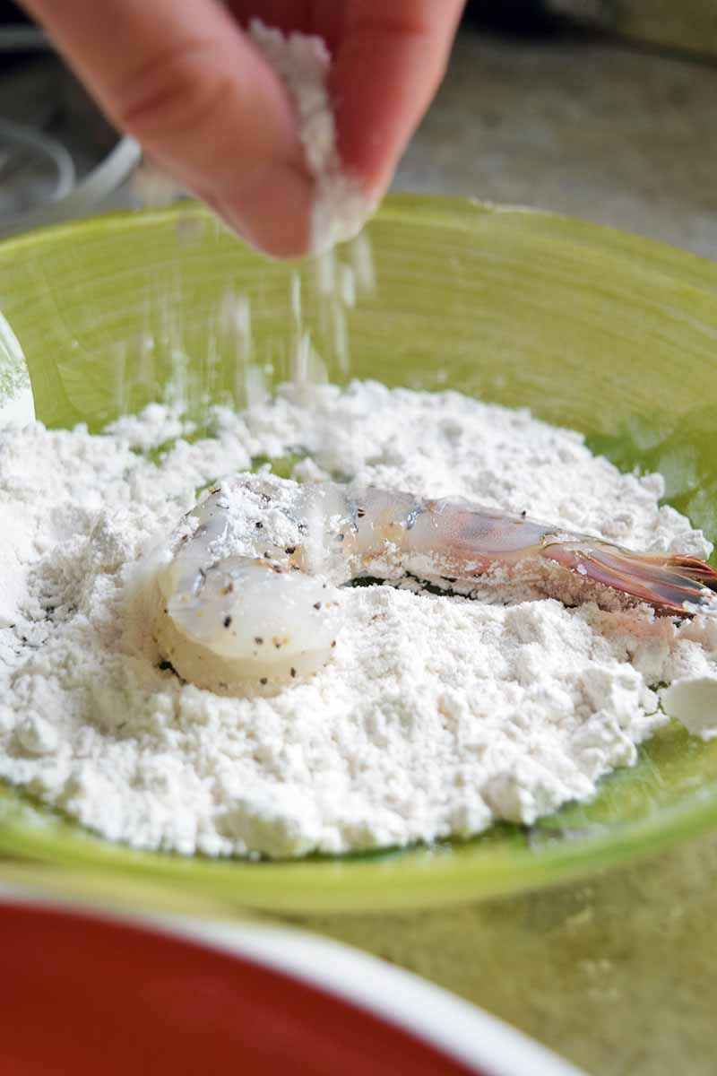Vertical image of a hand sprinkling flour onto a raw shrimp that has been seasoned with salt and pepper, in a shallow green glass bowl.