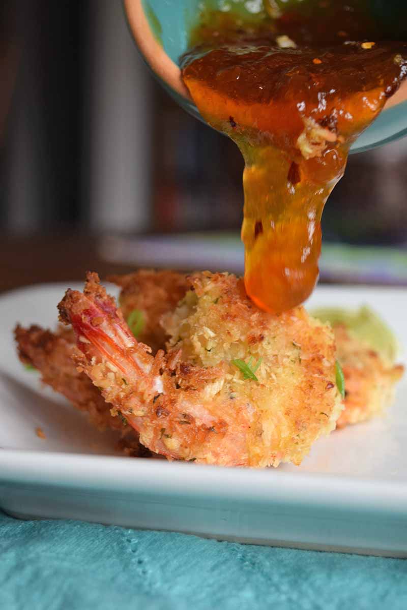 Vertical image of sweet and sour sauce being poured from a blue and terra cotta bowl onto baked coconut shrimp on a white plate, on top of a blue cloth.
