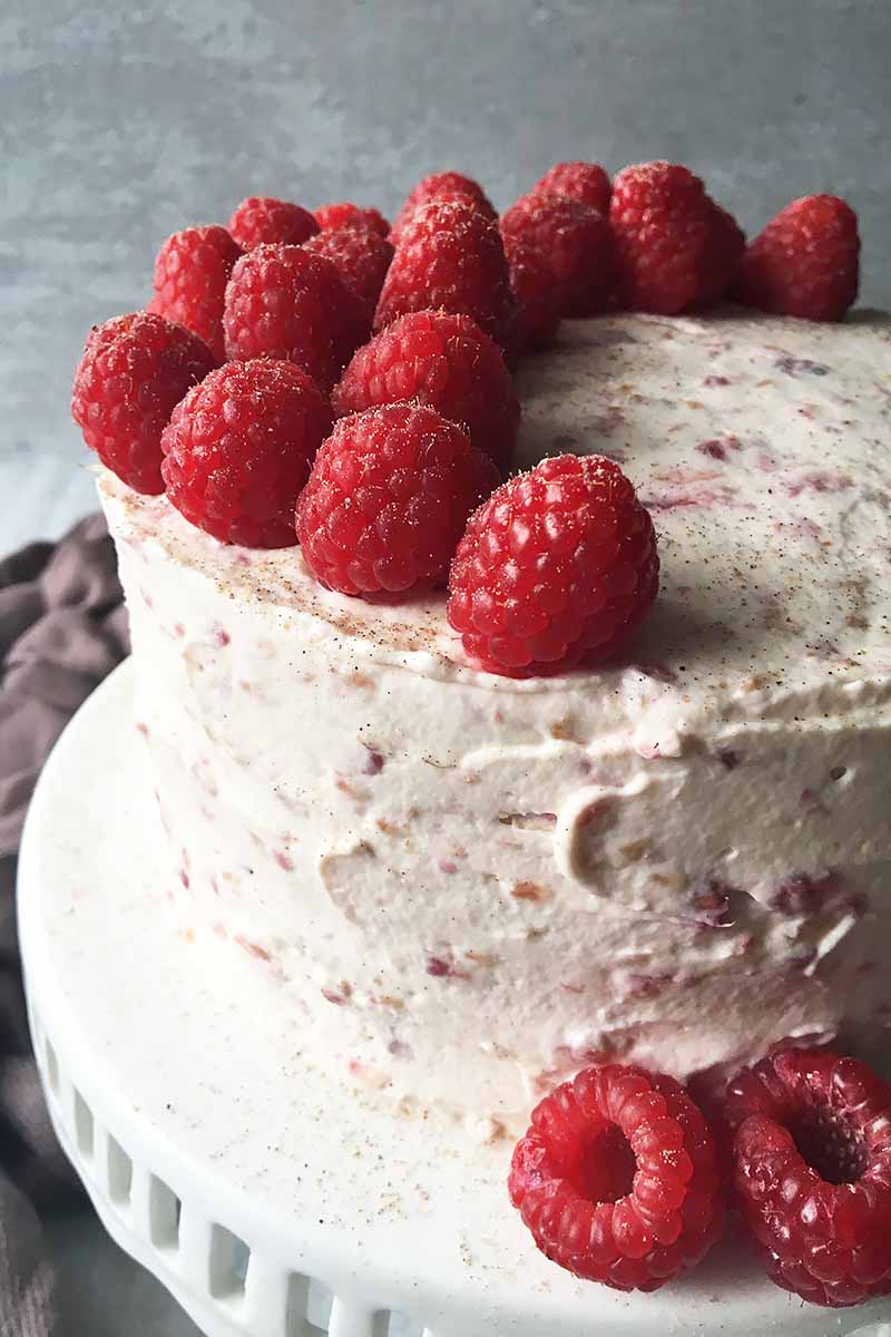 Vertical image of a whole decorated cake with raspberry frosting, and a garnish of more fresh whole raspberries.