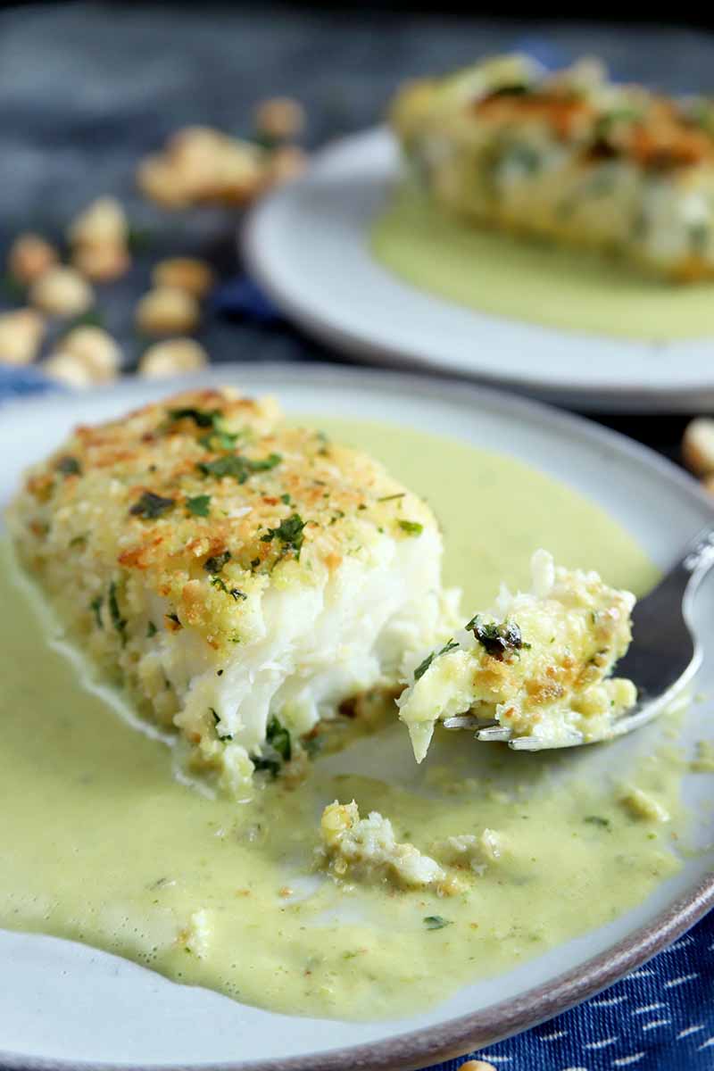 Vertical image of a white plate of nut-crusted halibut with coconut sauce, with a forkful of the dish and another identical plate in the background, on a blue-gray surface with scattered nuts in soft focus.