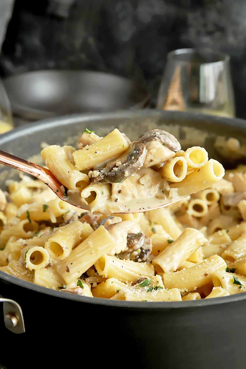 Vertical image of a spoon holding a pasta dish with herbs and mushrooms in a pot.