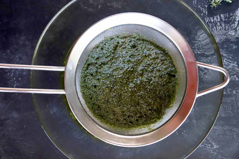 A green herbal mash in a mesh strainer set over a glass mixing bowl, on a gray slate surface.