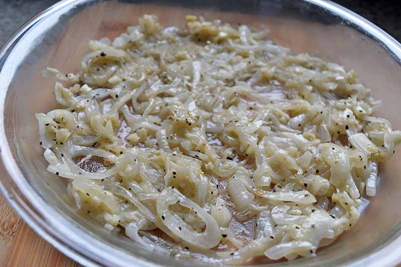 Horizontal image of sauteed sliced onions and minced garlic spread in an even layer in the bottom of a glass pie dish, on a light brown countertop.