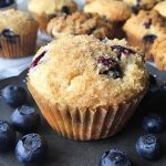 Horizontal image of blueberry muffins on a dark pan with fresh fruit surrounding them.