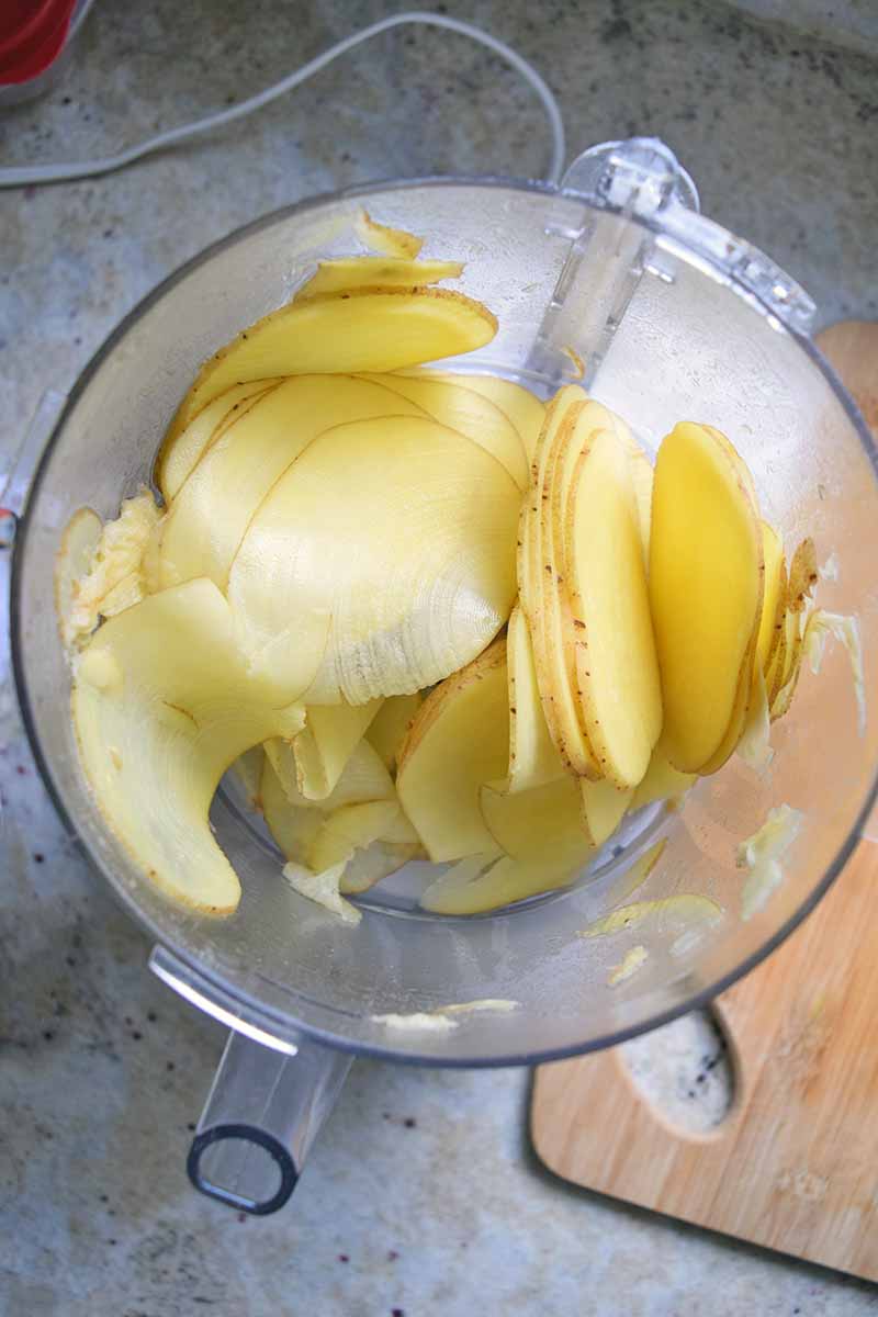 Vertical overhead image of thinly sliced potato in the plastic canister of a food processor, on a gray countertop with a wooden cutting board.