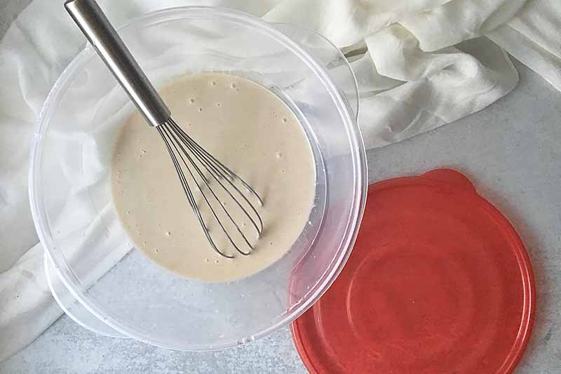 Horizontal image of a whisk and a white liquid in a plastic container with a red lid on a white towel.