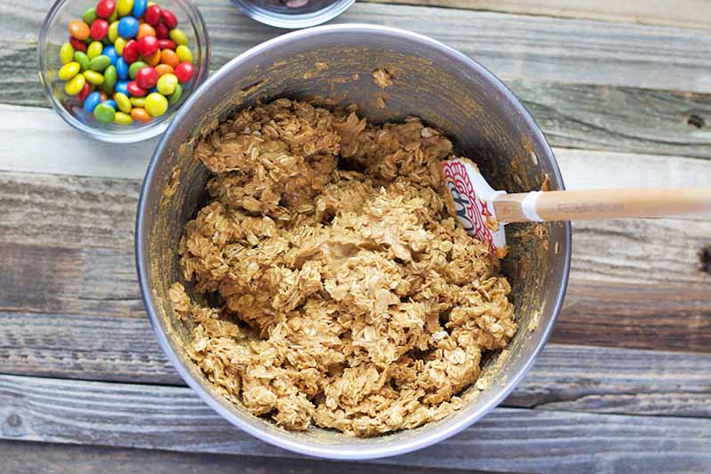 Horizontal image of a stainless steel bowl containing an oat batter mixture being stirred with a white rubber spatula with a wooden handle, on a wood surface with a small bowl of multicolored M&Ms.