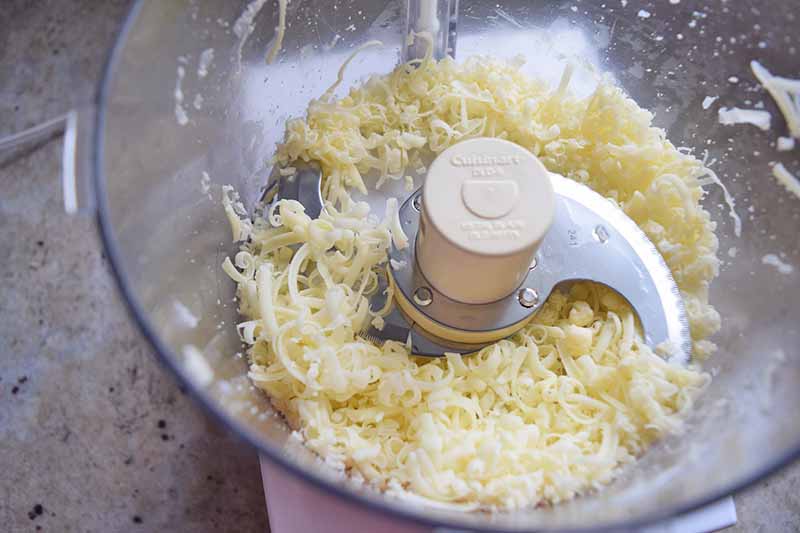 Horizontal closely cropped image of shredded cheese in the bottom of a food processor on a gray speckled countertop.
