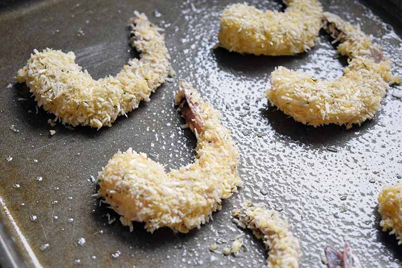 Horizontal image of coconut and panko breaded shrimp with the tails on, arranged in two rows on a metal baking sheet that has been sprayed with oil.