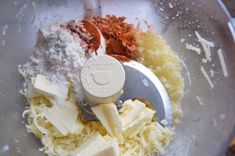 Horizontal overhead image of shredded cheese, butter, paprika, and flour in a food processor.
