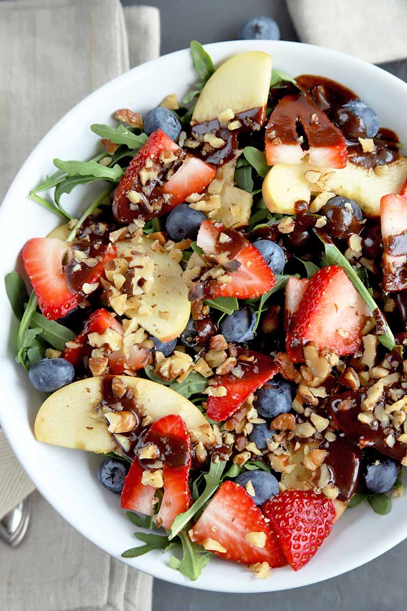 Overhead vertical closely cropped image of a shallow white bowl of salad with arugula, strawberries, blueberries, apple slices, chopped walnuts, and chocolate dressing, on a gray surface with a folded beige cloth napkin and a fork to the left.
