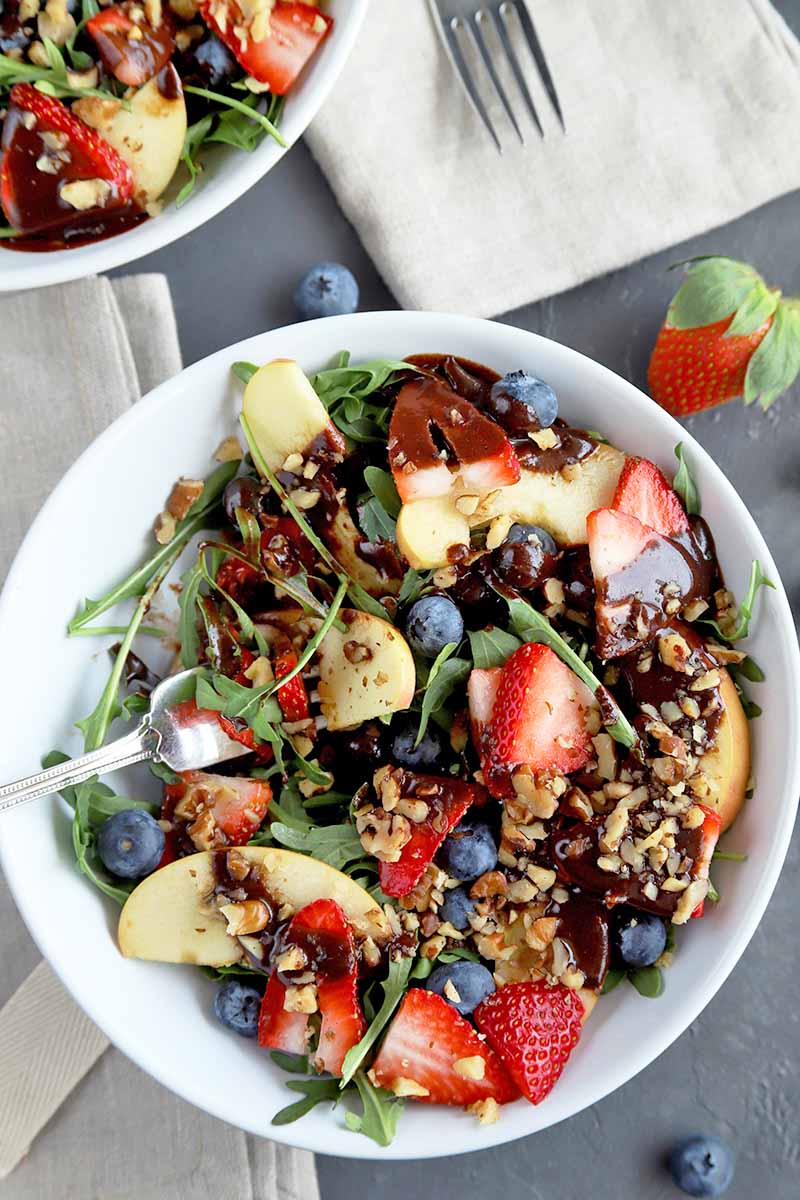 Overhead image of a shallow white ceramic bowl of fruit, nut, and arugula salad topped with chocolate balsamic dressing, on a gray surface with two folded beige cloth napkins, a fork, scattered blueberries, a whole strawberry, and another identical bowl in the top left corner of the frame.