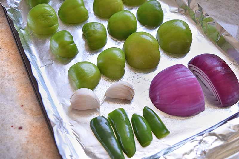 Horizontal image of a baking sheet covered with foil and topped with halved tomatillos, serrano peppers, and a red onion arranged in rows, alongside two whole cloves of garlic, on a kitchen countertop.