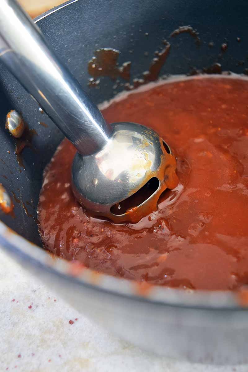 An immersion blender purees a red sauce in a nonstick cooking pot, on a speckled gray surface.