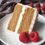 Horizontal image of a two-layer spelt cake with a white filling and light pink frosting on a white plate garnished with raspberries.