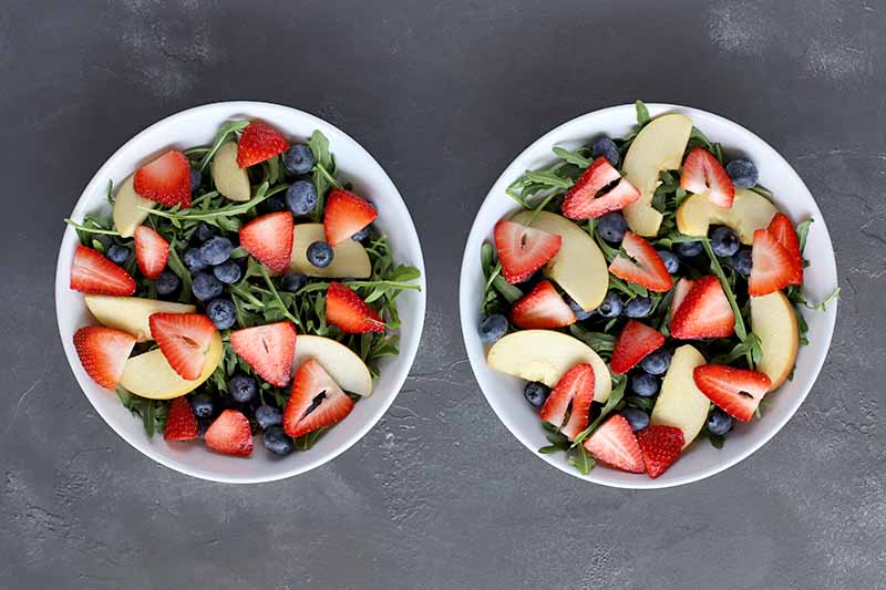 Vertical overhead image of two white ceramic bowls filled with arugula topped with fresh berries and apple slices, on a gray surface.