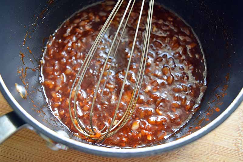 A wire whisk stirs a tomato-based mixture in a nonstick pan, on a beige wood background.