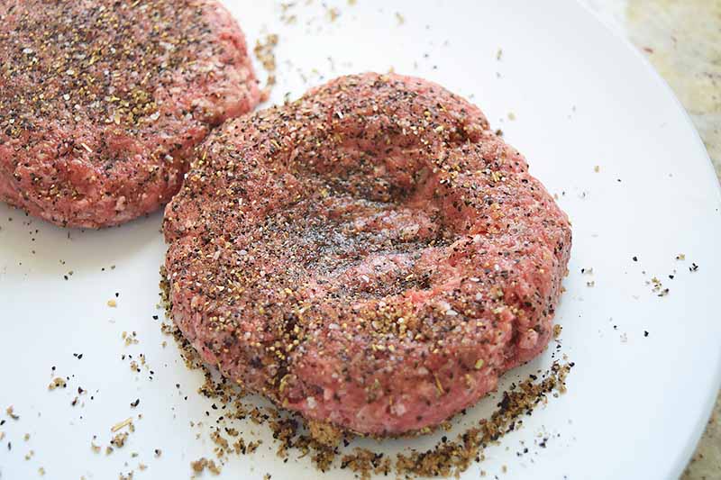 Horizontal image of two ground beef burger patties coated in a spice rub, with indentations made in the centers, on a white plate.