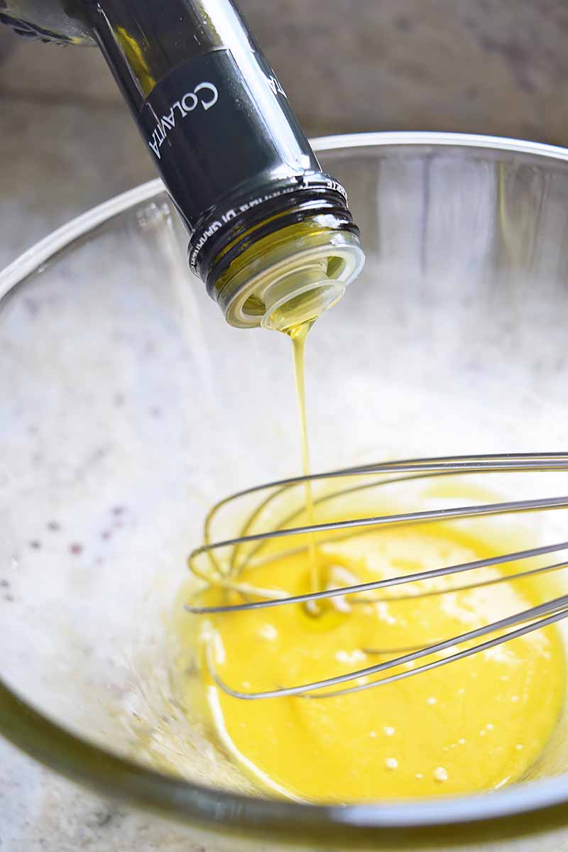 Closely cropped vertical image of olive oil being poured from a bottle into a mixing bowl with a yellow mixture at the bottom, being stirred with a wire whisk.