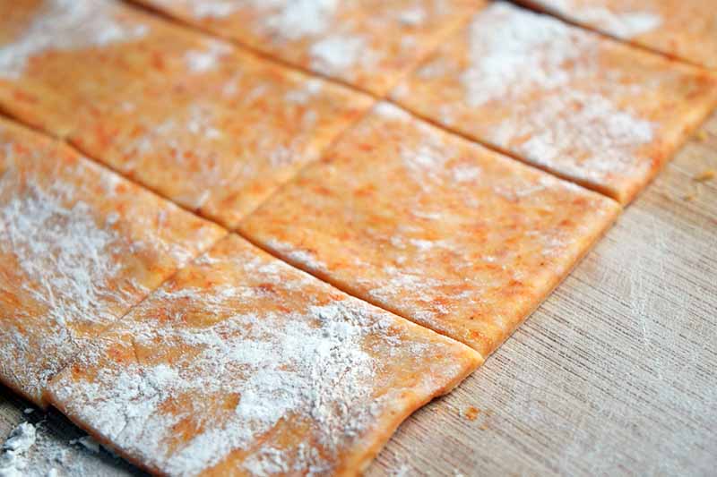 Horizontal closeup image of orange dough rolled out thin and scored to be portioned into square cracker shapes, on a countertop lightly dusted with flour.
