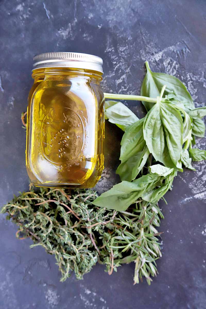 Overhead vertical image of a glass jar of golden yellow olive oil with a metal lid, resting on its side on a gray slate surface, with sprigs of fresh thyme and basil.