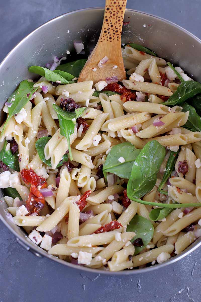 Vertical closely cropped overhead image of cooked pasta with crumbled feta, baby spinach, and chopped sun-dried tomatoes in a large stainless steel bowl with a wooden spoon, on a gray surface.