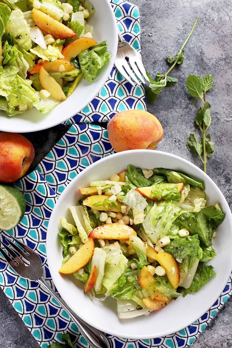 Vertical image of two white bowls filled with a stone fruit, fresh mint, and corn green salad on a blue towel next to whole stone fruit and whole mint sprigs.