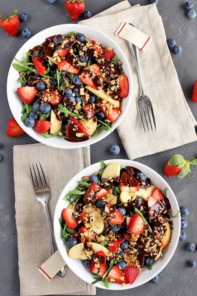 Overhead image of two white ceramic bowls filled with a homemade salad made with arugula, fresh fruit, and chopped nuts, topped with homemade balsamic chocolate dressing, on a gray surface with scattered berries and folded beige cloth napkins topped with forks.