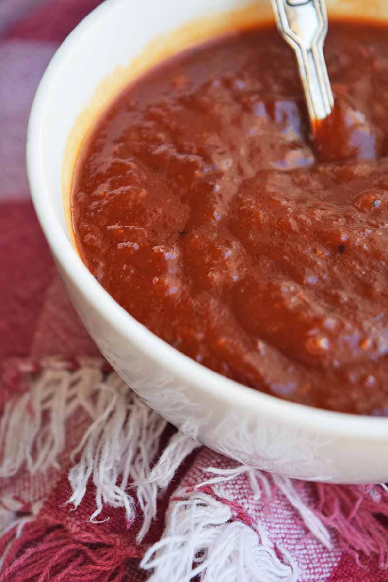 Vertical closely cropped image of a white bowl of red barbecue sauce with a spoon in the bowl, on a red and white checkered cloth with fringe.