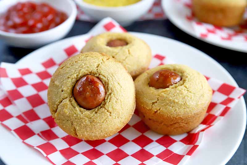 Horizontal closeup image of three corn dog muffins arranged on a white plate on top of a red and white paper liner, with another plate of the same and two small white dishes of ketchup and mustard in the background, on a dark gray stone surface.