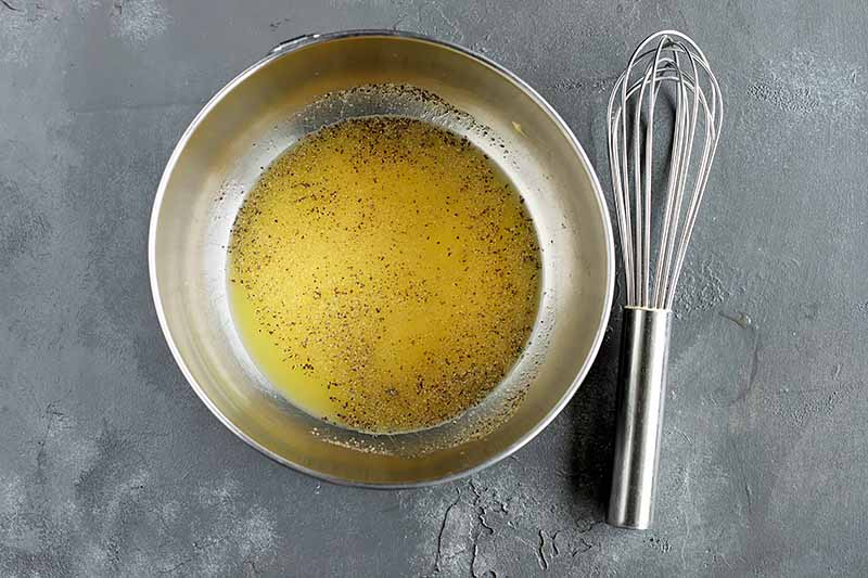 Horizontal image of a metal bowl with yellow dressing next to a whisk.