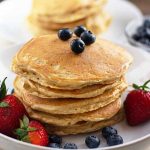 Horizontal image of a stack of flapjacks on a white dish with fresh strawberries and blueberries, with a bowl of blueberries and syrup in the background.